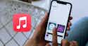 Apple Music Debuts Lyrics Translation and Karaoke Features meta Hand holding iPhone displaying Apple Music library with album covers. Red music note app icon visible in foreground on tiled surface.