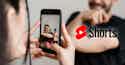 You Tube Shorts Everything You Need to Know meta Manos sosteniendo smartphone grabando a hombre barbudo, logo rojo y negro de YouTube Shorts superpuesto en la imagen.